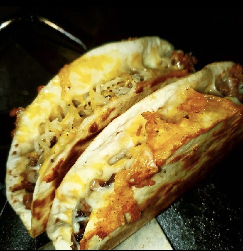 Two cheesy brisket tacos crisped on a dark flat-top surface