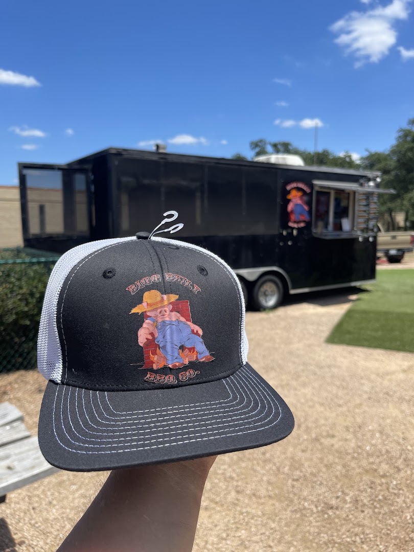 A Bigg Belly BBQ Co. branded trucker hat held up in front of the pit