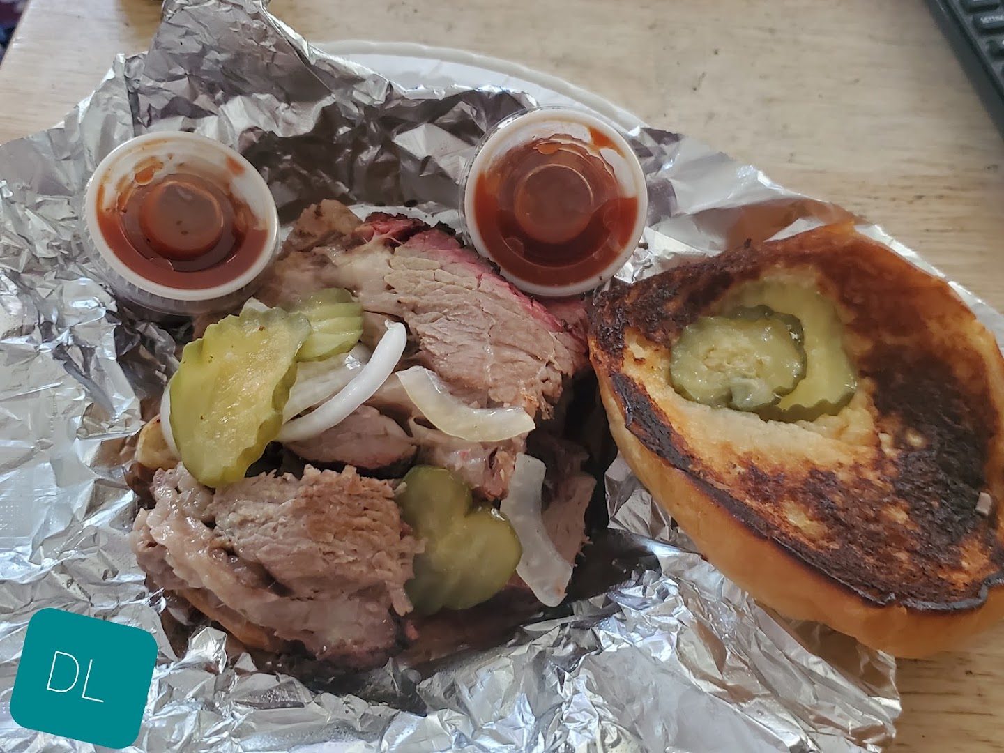 Open brisket sandwich on foil with pickles, onion and two cups of barbecue sauce